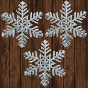 以下が含まれることがあります： 茶色の木製の背景に、銀色のグリッターで飾られた雪の結晶が3つあります。雪の結晶は三角形の形に配置されており、下の雪の結晶は下向きになっています。