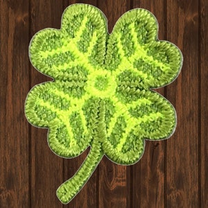 May include: A green four-leaf clover embroidered patch with a yellow outline. The clover is on a brown wooden background.