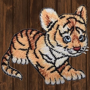 May include: A small, embroidered tiger cub with orange and black stripes, white accents, and blue eyes. The tiger cub is lying on its belly with its head turned to the side.