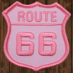 Puede incluir: Parche rosa Route 66 con fondo blanco y forma de escudo. El texto "ROUTE" está por encima del número "66".