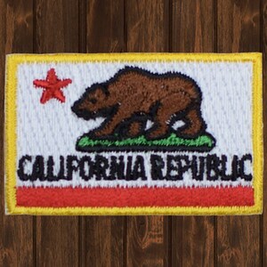 Könnte beinhalten: Ein weißer gestickter Aufnäher mit einem roten Stern, einem braunen Bären und grünem Gras. Der Text "CALIFORNIA REPUBLIC" ist in Schwarz unter dem Bären gestickt. Der Aufnäher hat einen gelben Rand.