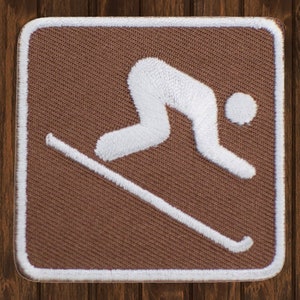 May include: Brown fabric patch with a white embroidered skier silhouette. The skier is in a downhill position with skis and poles.