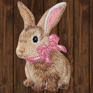 May include: Embroidered patch of a brown bunny with a pink bow and pink inner ears. The bunny is facing left, with detailed stitching creating a textured appearance. The patch is likely for clothing or accessories.