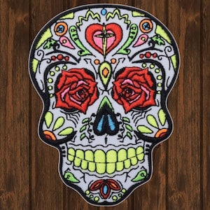 May include: A colorful embroidered sugar skull patch with red roses, a green heart with a cross, and a white background.