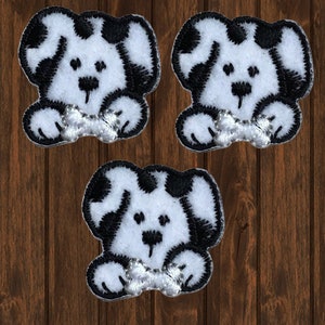 May include: Three embroidered patches featuring a cartoon dog design. Each patch has a white body with black spots and black outlines. The dog is holding a silver bone. The patches are on a brown wooden background.
