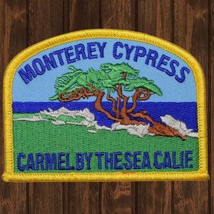 Op de afbeelding: Een geborduurd geel patch met een blauwe achtergrond met een gestileerde Monterey Cypress-boom met een groene basis en blauw water. De tekst "Monterey Cypress" is in blauw en de tekst "Carmel by the Sea Calif" is in blauw.