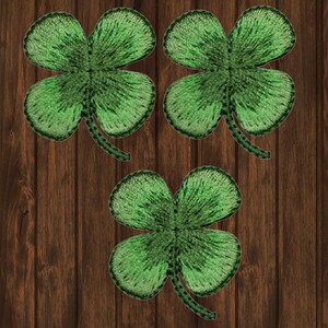 May include: Three embroidered green four-leaf clovers on a dark brown wooden background. The clovers have detailed stitching and a textured appearance, with varying shades of green. The image is a close-up, highlighting the clovers' details.