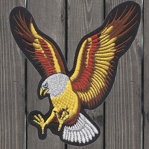 Peut inclure: Un écusson brodé représentant un aigle jaune, rouge et marron aux ailes déployées. L'aigle est tourné vers la gauche et a une tête et un bec blancs.