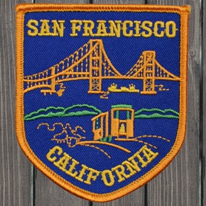 Puede incluir: Un parche bordado azul y naranja con el texto "SAN FRANCISCO CALIFORNIA" y una imagen del Puente Golden Gate y un tranvía.