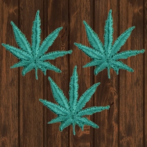 May include: Three teal green embroidered marijuana leaf patches. The patches are on a brown wood background.