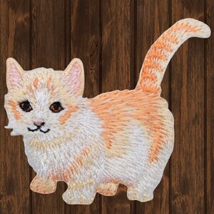 May include: A white and orange embroidered patch of a small, short-legged cat with a curled tail.