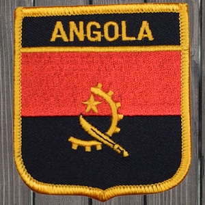 May include: A red, black, and gold embroidered patch with the word "ANGOLA" in yellow lettering at the top and the Angolan flag symbol in the center.