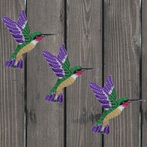 Könnte beinhalten: Drei gestickte Kolibri-Aufnäher mit violetten, grünen und goldenen Akzenten. Die Kolibris sind im Flug mit ausgebreiteten Flügeln.