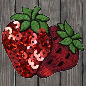 May include: Embroidered strawberry patch with red sequins and green leaves. One strawberry is covered in sequins, the other is embroidered. The patch is on a wooden background.