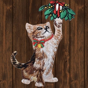 May include: An embroidered iron-on patch of a brown and white tabby cat reaching up to grab a sprig of mistletoe with red berries and a red bow. The cat is wearing a red collar with a green bow and a gold bell.
