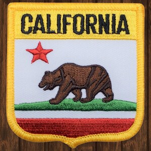 Puede incluir: Un parche bordado amarillo con las palabras "CALIFORNIA" en letras negras. El parche presenta una estrella roja, un oso marrón caminando sobre un campo verde y una franja roja, blanca y verde.