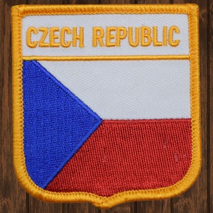 Peut inclure: Un écusson blanc avec un triangle bleu et une bande rouge, avec les mots "CZECH REPUBLIC" en lettres jaunes en haut. L'écusson est entouré d'une bordure jaune.