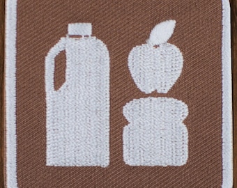 Könnte beinhalten: Weiß gesticktes Design einer Milchkanne, eines Apfels und eines Laibes Brot auf braunem Stoffhintergrund.