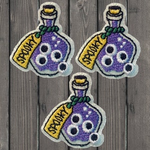 May include: Three purple glass bottles with a green stopper and a yellow tag that says "Spooky" on it. Each bottle has three white eyeballs inside.