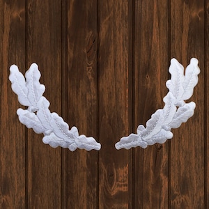 Può includere: Due corone di foglie di quercia ricamate bianche su uno sfondo di legno marrone.