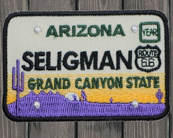 Puede incluir: Un parche bordado blanco y verde con el texto "Arizona" y "Seligman" en negro. El parche también presenta un letrero verde de la Ruta 66 y el texto "Grand Canyon State". El parche tiene un paisaje desértico morado con dos cactus y una puesta de sol amarilla.