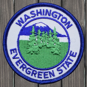 Puede incluir: Un parche bordado blanco con un borde azul. El parche presenta una cordillera con un pico nevado, un campo verde y una línea de árboles de hoja perenne. El texto "WASHINGTON EVERGREEN STATE" está escrito en un círculo alrededor de la imagen.