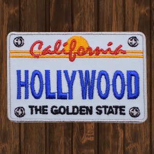 Könnte beinhalten: Weißer gestickter Patch mit einem roten und gelben Design einer kalifornischen Nummernschild. Das Schild zeigt "California" mit einer gelben Sonne und "HOLLYWOOD" in blauer Schrift. Der untere Teil des Schildes zeigt "THE GOLDEN STATE" in schwarzer Schrift.