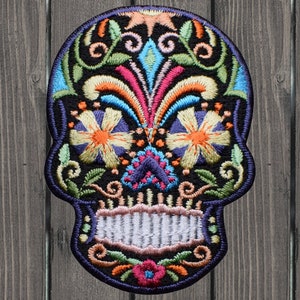 May include: A colorful embroidered sugar skull patch with intricate floral designs in shades of orange, yellow, green, blue, pink, and purple. The skull is outlined in black and has a white smile.