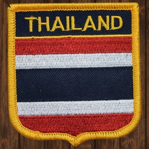 May include: A red, white, and blue embroidered patch with the word "THAILAND" in yellow lettering. The patch is surrounded by a yellow border and has a slightly curved bottom edge.
