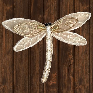 May include: A beige fabric dragonfly with beaded accents. The dragonfly is on a brown wooden background.
