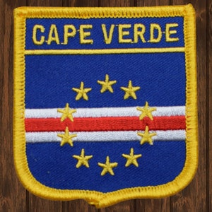 May include: A blue and white embroidered patch with a yellow border. The patch features the flag of Cape Verde with a red and white stripe and nine yellow stars. The text "CAPE VERDE" is embroidered in yellow at the top of the patch.