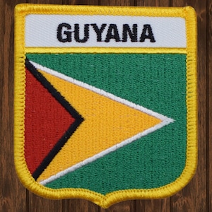 May include: Embroidered patch featuring the flag of Guyana. The patch has a shield shape with a yellow border. The flag design includes green, yellow, red, black, and white colors, with the word "GUYANA" at the top.