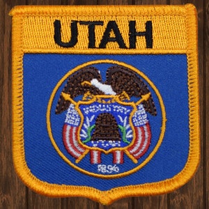 May include: A blue and gold embroidered patch with the word "UTAH" in black lettering at the top and the state seal of Utah in the center. The seal features a bald eagle with wings spread, holding a shield with a beehive and the words "Industry" and "State of Utah".