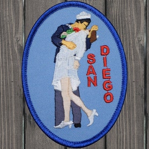 Op de afbeelding: Geborduurde patch met een matroos die een vrouw kust, met de tekst "SAN DIEGO" in het rood. De patch heeft een ovale vorm met een blauwe achtergrond en een donkerblauwe rand, die een romantische scène uitbeeldt.