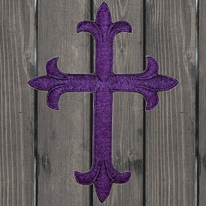 May include: A purple embroidered cross with a fleur-de-lis design. The cross is against a woodgrain background.
