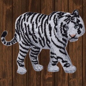 May include: Embroidered patch of a white tiger with black stripes, walking from left to right. The tiger has a pink nose and blue eyes. The patch is on a brown wooden background.