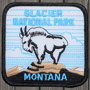 May include: A white embroidered patch with a black outline. The patch features a mountain goat standing on a rocky outcrop. The text "GLACIER NATIONAL PARK" is above the goat and "MONTANA" is below. The patch has a blue and white striped background.