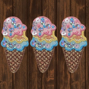May include: Three embroidered ice cream cone patches with pink, yellow, and blue ice cream and brown waffle cones. The patches are decorated with sequins.