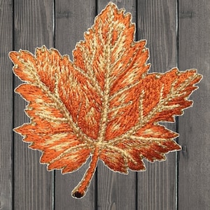 May include: A large, orange and gold embroidered maple leaf with a detailed vein pattern. The leaf is on a wood background.