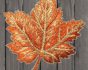 Può includere: Una grande foglia d'acero ricamata in arancio e oro con un motivo di venature dettagliato. La foglia è su uno sfondo di legno.