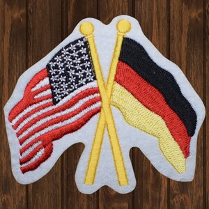 May include: A white felt patch with two crossed wooden sticks holding an American flag and a German flag. The American flag is red, white, and blue with stars. The German flag is black, red, and yellow.