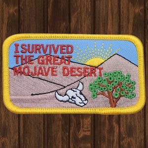 Può includere: Una toppa ricamata gialla con il testo "I Survived The Great Mojave Desert" in rosso e bianco. La toppa presenta un paesaggio desertico con un sole, un albero e un teschio.