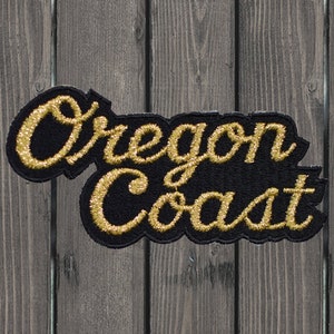 May include: Black embroidered patch with the words "Oregon Coast" in gold script lettering. The patch has a black border and is set against a wooden background.