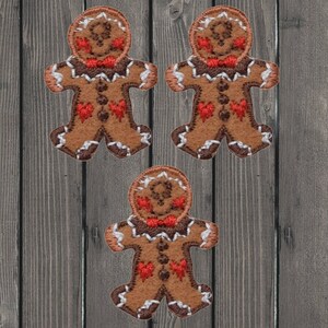 Könnte beinhalten: Drei Lebkuchenmann-Patches. Jeder Patch ist braun mit weißen und roten Akzenten. Die Lebkuchenmänner haben rote herzförmige Knöpfe und Fliegen. Die Patches sind mit weißen Nähten an den Rändern und Details bestickt.
