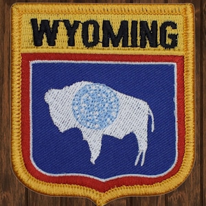 Op de afbeelding: Een geborduurde patch in geel en goud met het woord "WYOMING" in zwarte letters. De patch heeft een blauwe achtergrond met een wit silhouet van een buffel met een blauwe cirkel in het midden.