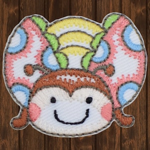Peut inclure: Un écusson brodé coloré à repasser représentant un papillon de dessin animé souriant avec des accents roses, bleus, jaunes et verts.