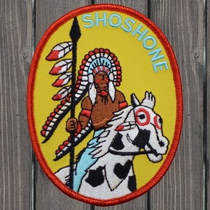 May include: An embroidered patch featuring a Native American warrior riding a horse, with the word "SHOSHONE" in blue lettering. The patch is yellow and red.