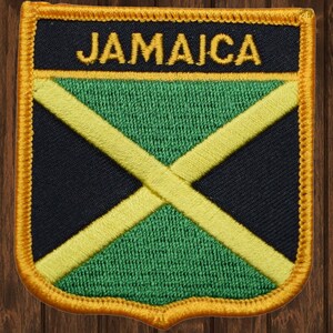 Puede incluir: Un parche bordado con la bandera de Jamaica, con la palabra "JAMAICA" en letras amarillas sobre un fondo negro. El parche está rodeado por un borde dorado.