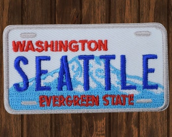May include: Embroidered patch in the shape of a Washington state license plate. The patch features the words "WASHINGTON" and "EVERGREEN STATE" in red, and "SEATTLE" in blue, with a blue mountain design.