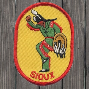 May include: A yellow oval embroidered patch with a red border. The patch features a green figure in a dancing pose holding a brown drum. The figure has a red and white feathered headdress. The text "SIOUX" is embroidered in red at the bottom of the patch.